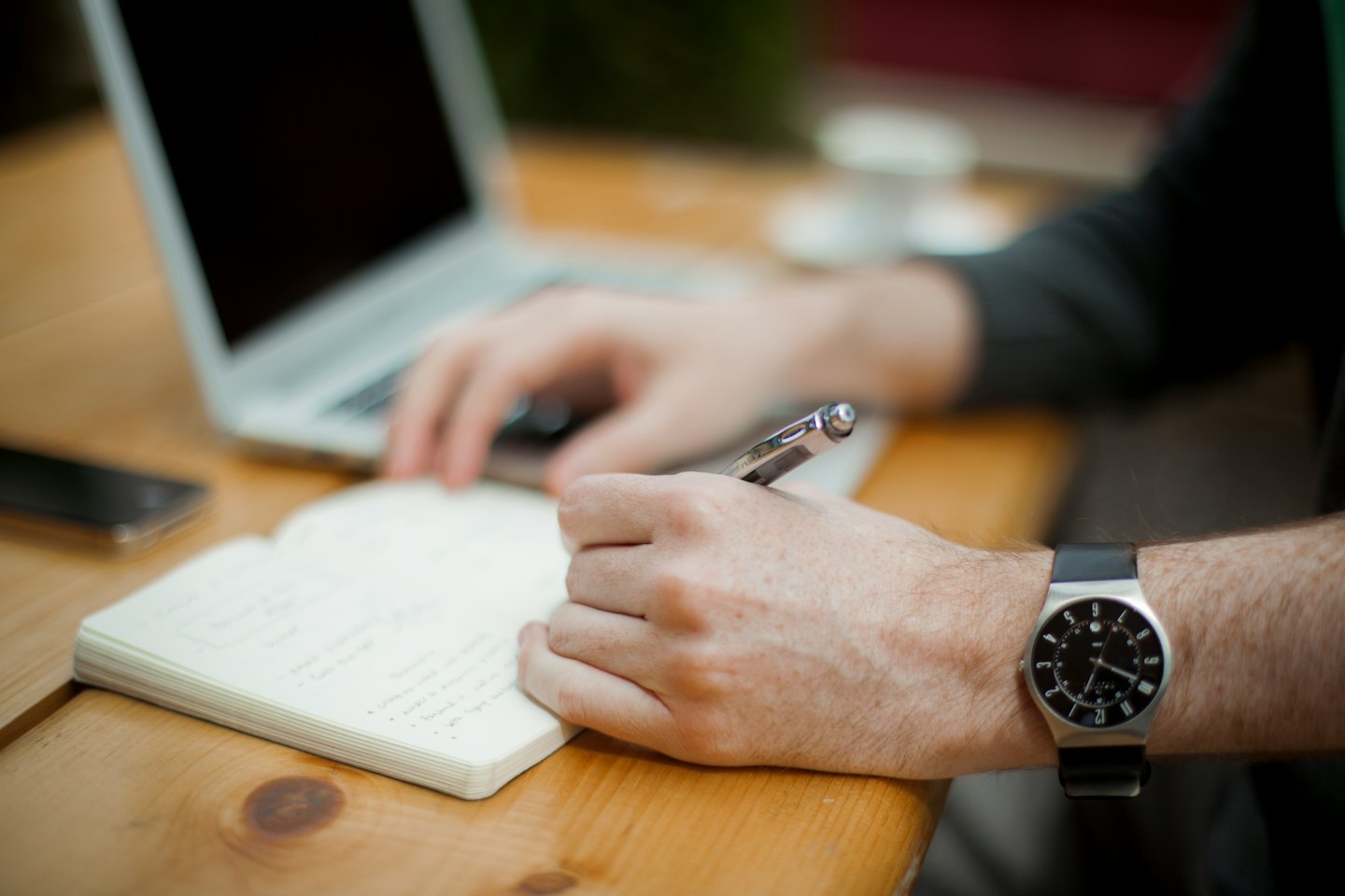Hands writing in a notebook next to a laptop.